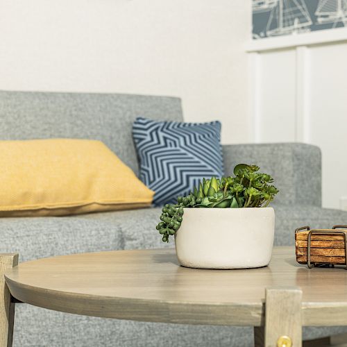 A cozy living room with a gray sofa, yellow and blue pillows, a round wooden table, a plant in a white pot, and a small wooden decor piece.