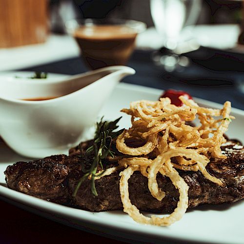 The image shows a dish with a steak topped with crispy onion strings, garnished with herbs, and accompanied by a side of sauce in a small gravy boat.