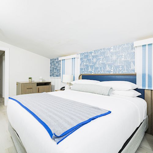 A cozy bedroom with a large bed, blue and white decor, wooden furniture, and a wallpapered accent wall. The room is bright and inviting.