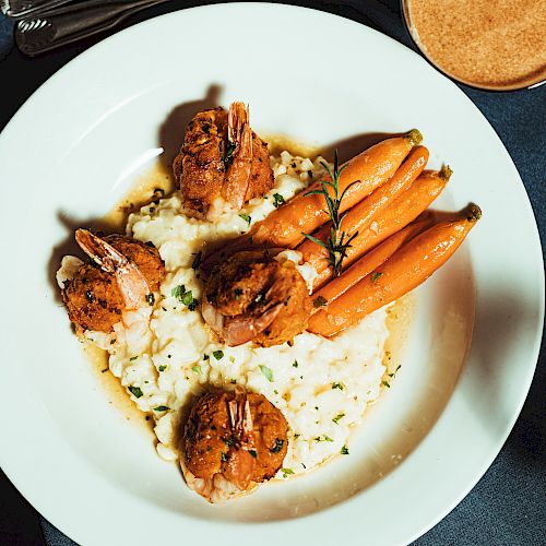 A plate of shrimp on mashed potatoes with glazed carrots and a sprig of rosemary, placed on a table with a drink.