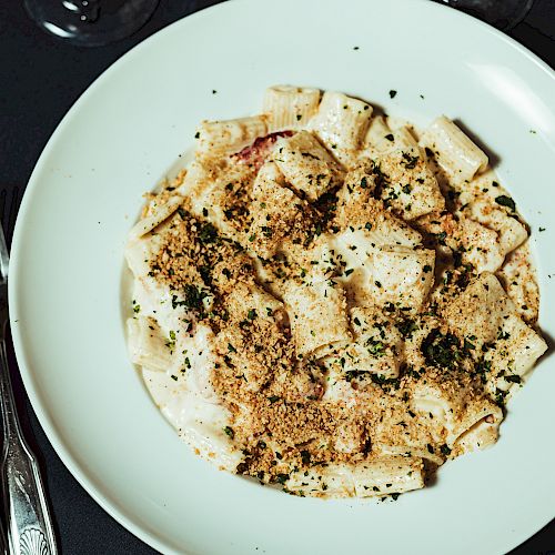 A plate of pasta with a creamy sauce, topped with breadcrumbs and herbs, next to utensils and an empty plate on a dark tablecloth.