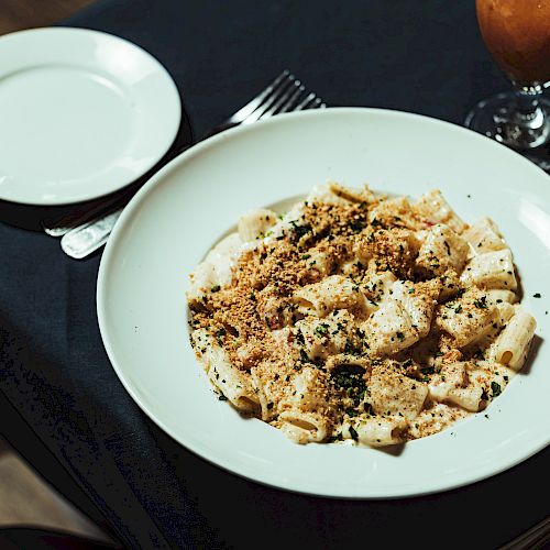 A white bowl of pasta with sauce and herbs on a table with a bread plate, fork, spoon, and drinks.