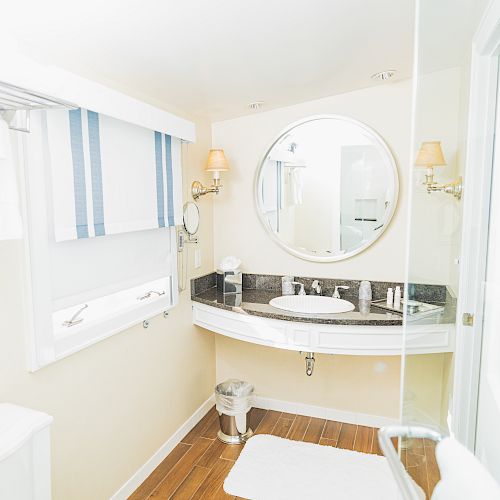 A well-lit bathroom featuring a round mirror above a sink, towel on a rack, wooden floor mat, and wall-mounted lights. The room is bright and clean.