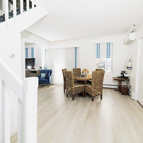 A bright living room with white walls, wooden floors, wicker furniture, and a staircase leading up.