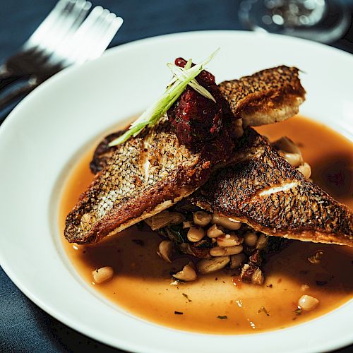 A white plate with seared fish fillets on a bed of beans and vegetables in a savory sauce, garnished with green onions, fork, and knife nearby.