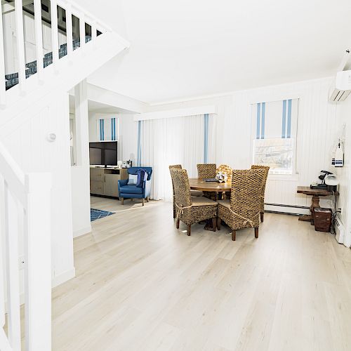 The image shows a bright, modern living space with light wood flooring, a staircase, wicker dining chairs, and a TV area with blue furniture.