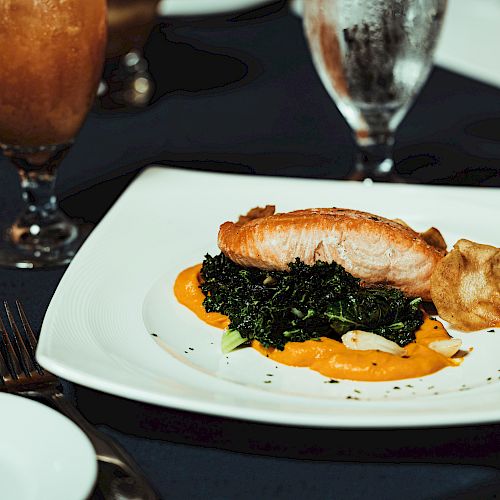 A dish featuring a piece of cooked salmon on a bed of greens and orange sauce, served with a drink and utensils on a table.