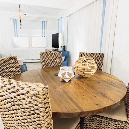 A dining area with a wooden table, wicker chairs, and decorative items including a geometric sculpture and wicker ball on the table, in a bright room.