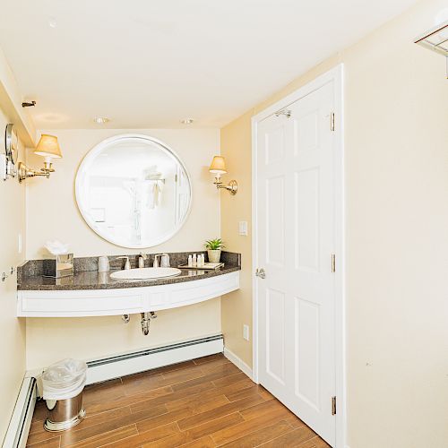 A bright bathroom with a round mirror, sink, towel rack, and toilet. There are also lights on either side of the mirror and a wooden floor.