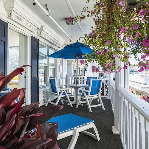 A cozy porch with seating, a blue umbrella, potted plants, and hanging flowers, overlooking a scenic view with a clear sky.