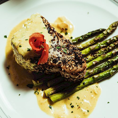 A plated dish featuring a seasoned piece of meat with a creamy sauce, garnished with herbs and red pepper, served atop a bed of grilled asparagus.