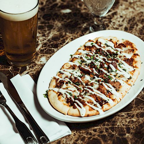 A flatbread pizza topped with sauce and herbs is on a white plate beside a glass of beer, with a knife, fork, and napkin nearby on a marble table.