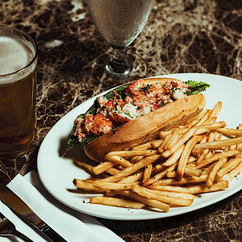 A plate of fries and a lobster roll on a marble table, accompanied by a glass of beer and water, with utensils and a napkin nearby.