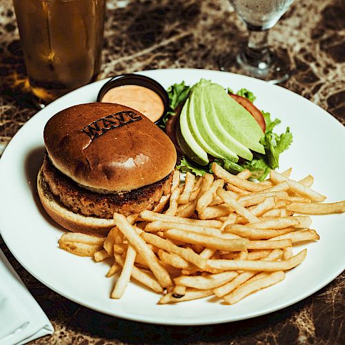 A plate with a burger, fries, avocado slices, and greens, accompanied by a drink and utensils on a marble table.