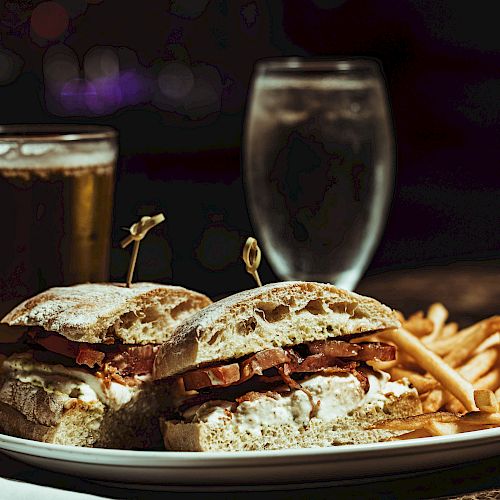 A plate with a sandwich and fries is in the foreground, accompanied by a glass of beer and a glass of water in the background.