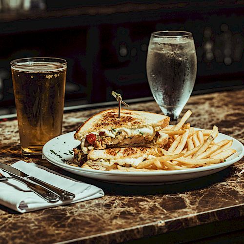 A sandwich, fries, a glass of beer, and a glass of water are on a marble table with utensils on a napkin.