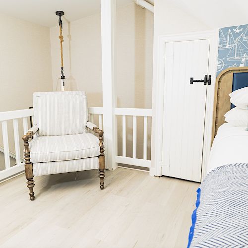 A cozy bedroom with a striped chair, a bed with blue and white linens, nautical-themed wallpaper, and white walls and flooring.