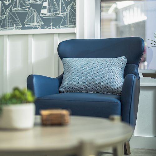 A cozy room with a blue armchair with a cushion, nautical-themed wall art, and a round table with a plant and small decor item in the foreground.