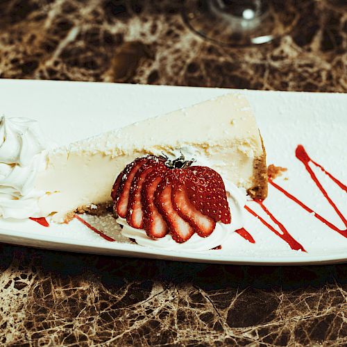 A slice of cheesecake garnished with sliced strawberries, whipped cream, and a drizzle of red sauce on a white plate sits on a marble-patterned table.