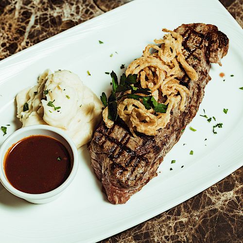 A plate with a grilled steak topped with fried onions, garnished with herbs, served with mashed potatoes, a small cup of sauce, and a glass of beer.