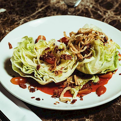 A wedge salad with iceberg lettuce, cherry tomatoes, crispy onions, bacon bits, and dressing, plated on a white dish over a marble table.