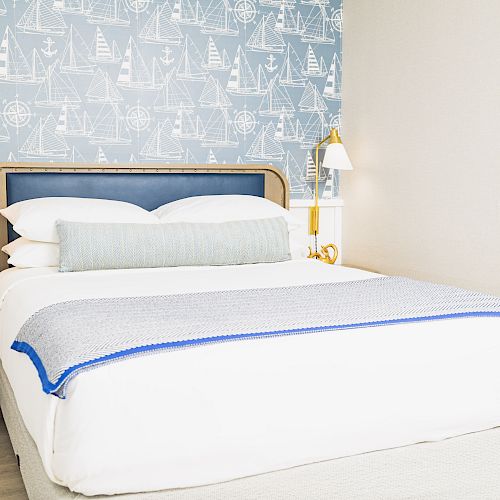 A well-decorated bedroom with nautical-themed wallpaper, a neatly made bed with white linens, a plaid throw, bedside tables, and wall-mounted lamps.