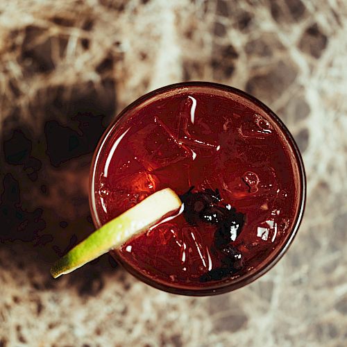 A top-down view of a red drink with ice, garnished with a lime slice and dark berries, on a textured surface.