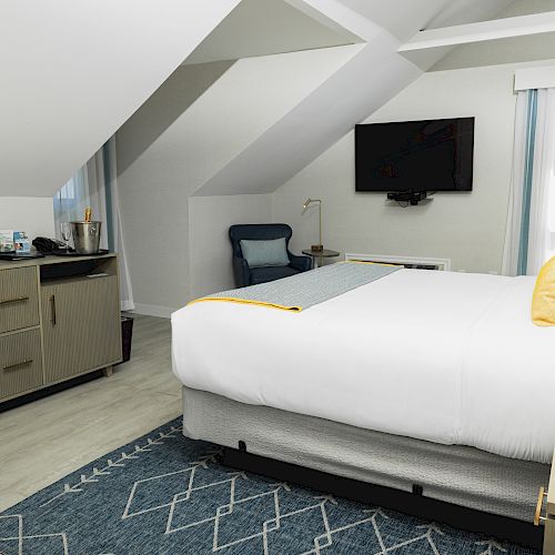 A modern bedroom under a sloped ceiling with a bed, TV, chair, and dresser. The dresser holds a coffee station and various items.