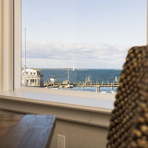 The image shows a view through a large window of a body of water with boats docked at a pier. There are two chairs in the foreground.