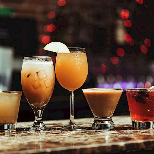 The image shows five different cocktails arranged on a bar counter, each garnished with fruit slices, and with a blurred background of lights.