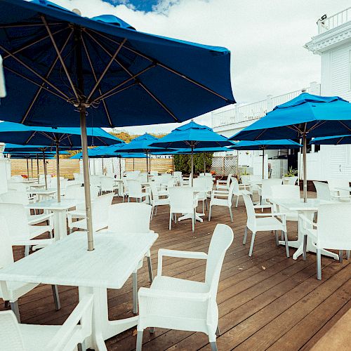 The image shows an outdoor patio with white tables and chairs, and blue umbrellas set up for a dining area, next to a white building.