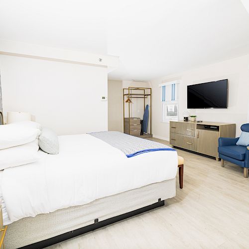 This image shows a modern hotel room with a white bed, blue chair, wall-mounted TV, dresser, and a clothing rack in a well-lit space.