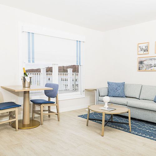 A cozy living room with a grey couch, blue pillows, a small round table, and a dining table with two chairs near a window.