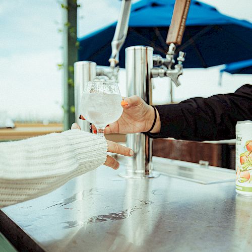 A person is handing a glass of a clear beverage to another person at an outdoor bar with blue umbrellas overhead, and a can of drink on the counter.