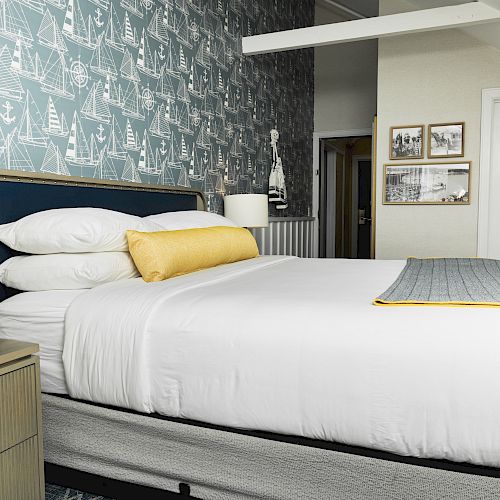 A neatly made bed with white linens and a yellow accent pillow in a well-decorated room featuring nautical-themed wallpaper and framed artwork.