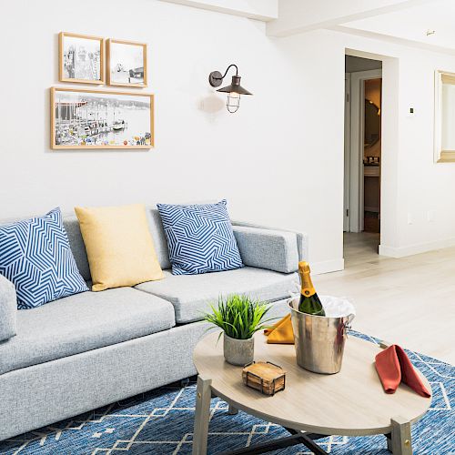 A cozy living room with a gray couch, blue-patterned pillows, wall art, a coffee table with items, and minimalistic décor.