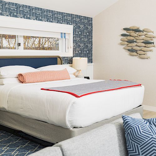 A cozy bedroom features a neatly made bed with a blue headboard, a nightstand, decorative pillows, wall art of fish, and a small window above the bed.