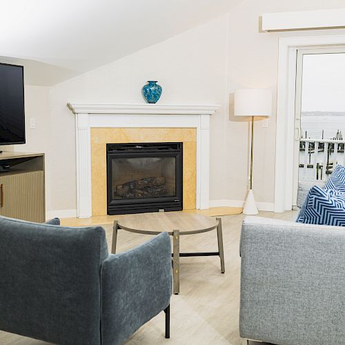 A cozy living room with a fireplace, TV, two chairs, a couch, a coffee table, and a lamp. There’s a view of a marina through the window.