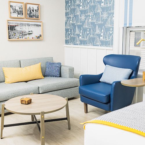 A cozy living room with a gray sofa, blue armchair, round coffee table, and nautical-themed wallpaper. There are framed photos on the wall.