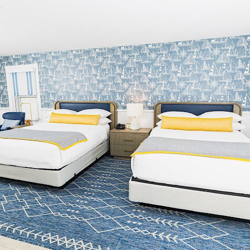 The image shows a bright hotel room with two double beds, blue and white bedding, a blue patterned carpet, and nautical-themed wallpaper.