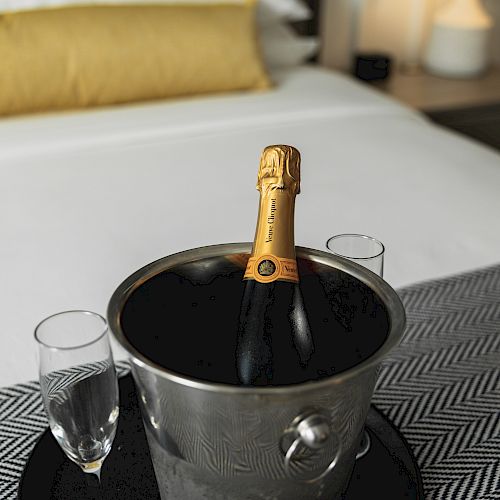 A bottle of champagne in an ice bucket with two glasses sits on a bed with a yellow pillow in the background.