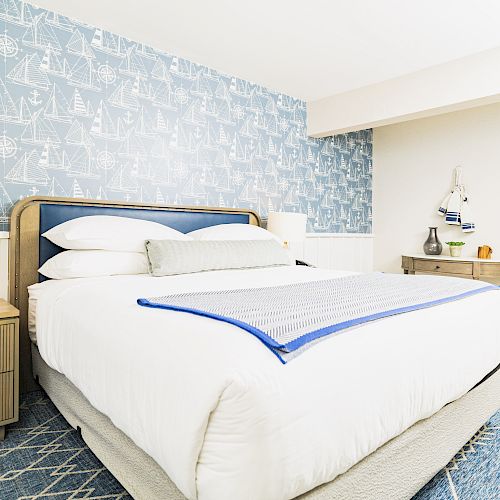 A bedroom with a nautical theme, featuring a patterned blue wallpaper, a bed with white linens, side table, and a console with accessories, ending the sentence.