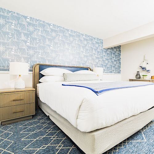 A cozy bedroom with a nautical theme featuring a bed, blue sailboat wallpaper, wooden nightstand with a lamp, and a dresser.