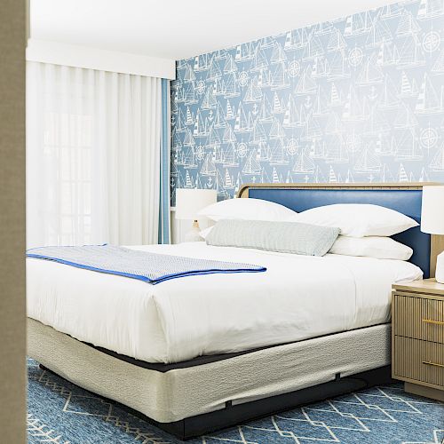 The image shows a neatly made bed in a modern bedroom with blue patterned wallpaper, a blue and white bedspread, and two bedside tables with lamps.