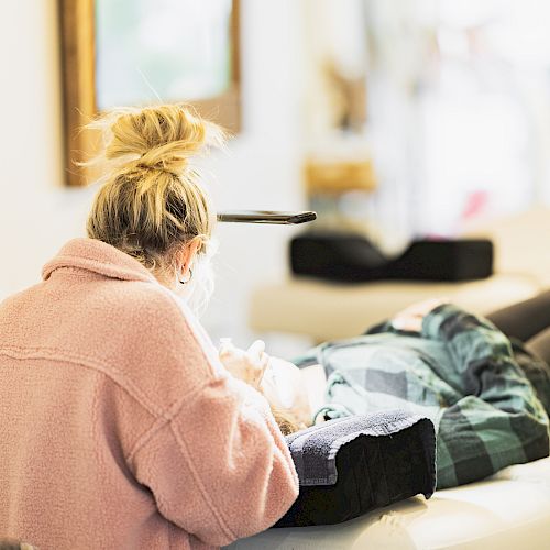 A person with a bun hairstyle, wearing a pink sweater, is attending to another person lying on a bed or chair in a well-lit, cozy room.