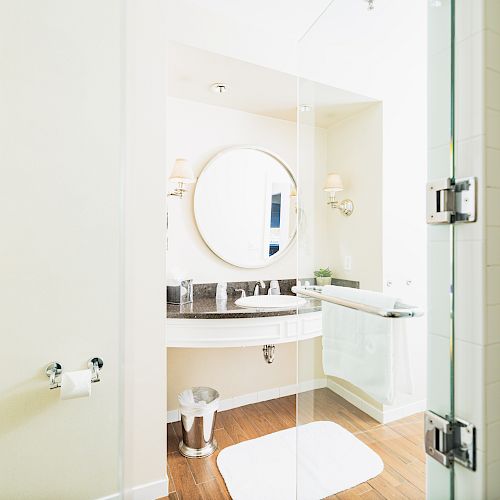 A modern bathroom with a round mirror, sink, towel rack, and toilet paper holder on the left wall; visible glass shower area.