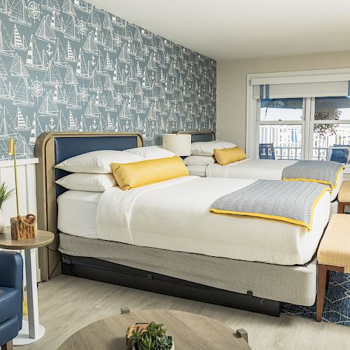 A neatly arranged hotel room with two beds, nautical-themed wallpaper, blue and yellow accents, and a cozy seating area by the window.