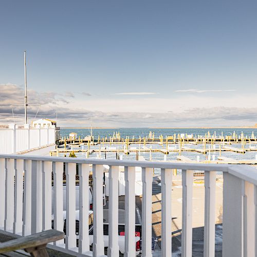 A calm seaside view shows a marina with empty berths, a white wooden railing in the foreground, and a coastal landscape under a clear sky.