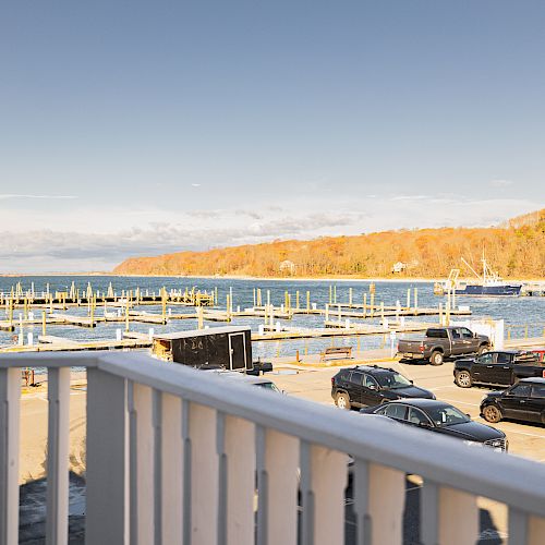 A scenic view of a lakeside marina with parked cars in the foreground, empty docks, and a tree-covered hill in the background under a clear sky.