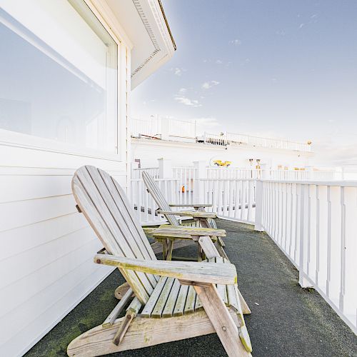 A sunlit balcony featuring weathered wooden chairs against a white railing and siding, under a clear blue sky with wispy clouds dotting the horizon.